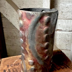 May include: A handmade, cylindrical ceramic vase with a textured surface. The vase features a blend of colours, including red, brown, and grey, with raised circular and curved decorative elements. The vase is displayed on a wooden surface.