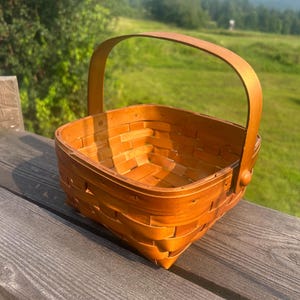 May include: A square, woven wooden basket with a curved handle. The basket is made of light brown wood and has a textured surface. The handle is attached to the sides with small wooden pegs. The basket is sitting on a wooden surface.