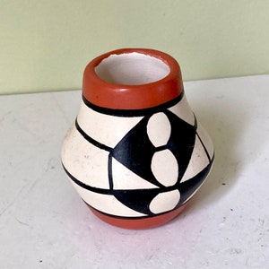 Puede incluir: Un pequeño jarrón de cerámica pintado a mano con un diseño geométrico en negro, blanco y rojo. El jarrón tiene un cuerpo redondeado y un borde ligeramente acampanado.