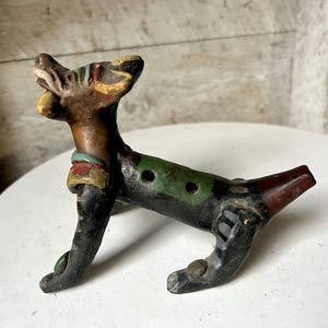 Puede incluir: Un silbato de cerámica con forma de perro pintado en marrón, negro, verde, rojo y amarillo. El silbato tiene cuatro agujeros en la parte superior.