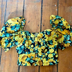 May include: A floral print top with short, puffed sleeves. The top features a black background with yellow roses and teal leaves. It has a square neckline and a button-down front. The fabric appears to be a lightweight material.