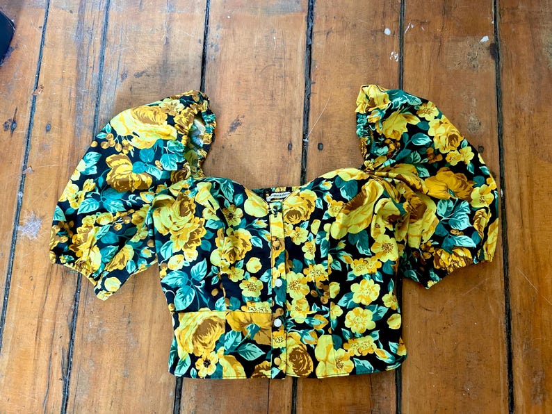 May include: A floral print top with short, puffed sleeves. The top features a black background with yellow roses and teal leaves. It has a square neckline and a button-down front. The fabric appears to be a lightweight material.