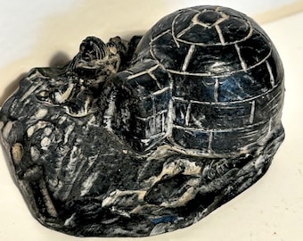 Può includere: Una scultura in pietra grigio scuro e nera che raffigura un igloo con una cupola arrotondata e motivi a blocchi. L'oggetto presenta una base strutturata con dettagli intagliati di una figura e del terreno circostante.