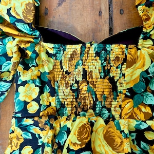 May include: A floral print dress featuring yellow roses and teal leaves on a black background. The bodice has a smocked design, and the sleeves are puffed. The dress is made of a lightweight fabric, perfect for a summer day.