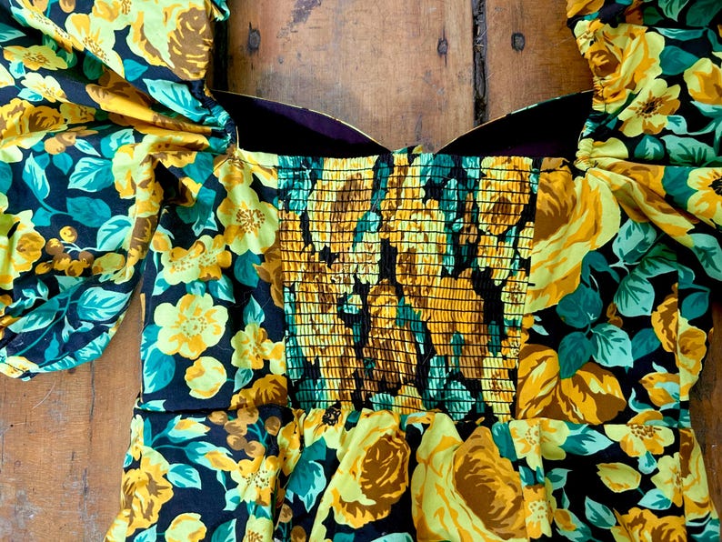May include: A floral print dress featuring yellow roses and teal leaves on a black background. The bodice has a smocked design, and the sleeves are puffed. The dress is made of a lightweight fabric, perfect for a summer day.