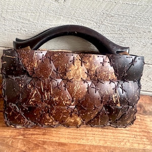 May include: A handcrafted handbag made from coconut shell pieces, stitched together with dark brown thread. The bag features a curved wooden handle and a geometric design. The bag is sitting on a wooden surface.