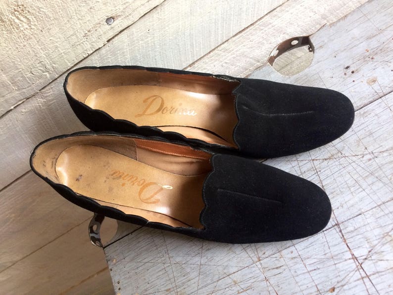 May include: A pair of black suede shoes with scalloped edges. The shoes have a pointed toe and a low heel. The brand name "Dorina" is visible on the inside of the shoes.