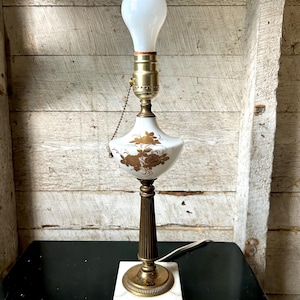 May include: Vintage table lamp with a white bulb, brass accents, and a white ceramic base with gold floral detailing. The lamp has a marble base and a ribbed brass column. The lamp is on a black surface.