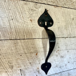 May include: Black, ornate door handle with a heart-shaped design. The handle is made of metal and has a keyhole. It is mounted on a weathered, white-painted wooden surface.