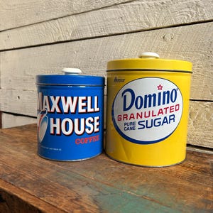 May include: Two vintage metal canisters. One is blue with "Maxwell House Coffee" text and a coffee cup graphic. The other is yellow with "Domino Granulated Pure Cane Sugar" text. Both have white lids.
