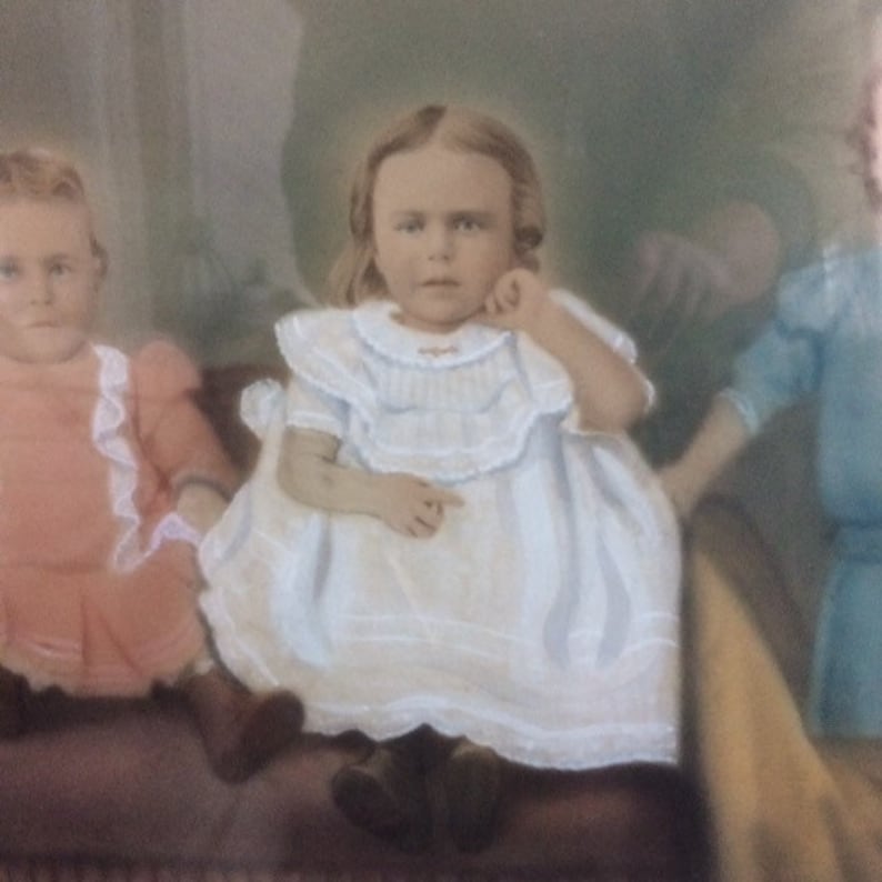 May include: Two young girls in vintage clothing pose for a portrait. The girl in the front is wearing a white dress with lace trim and has her hand on her chin. The girl in the back is wearing a pink dress with lace trim.