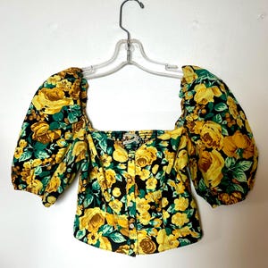 May include: A floral print top with a square neckline and short, puffed sleeves. The top features a black background with yellow roses and green leaves. It is buttoned down the front and hangs on a clear hanger.
