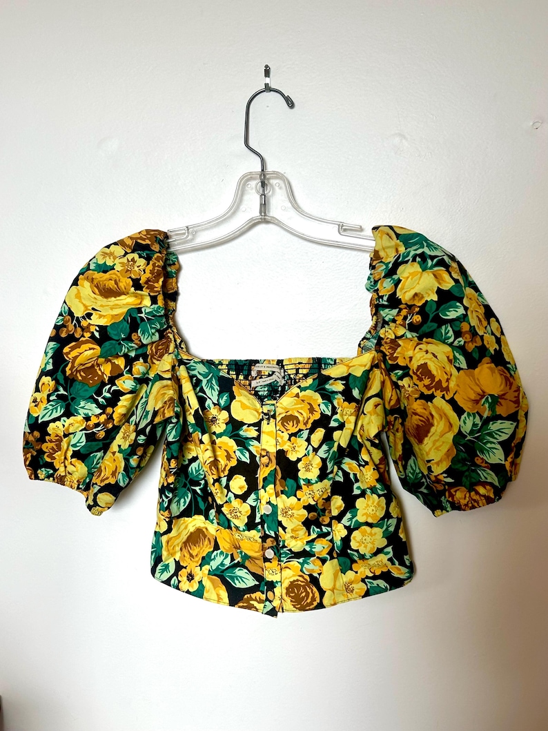 May include: A floral print top with a square neckline and short, puffed sleeves. The top features a black background with yellow roses and green leaves. It is buttoned down the front and hangs on a clear hanger.
