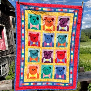 Puede incluir: Una colcha rectangular con una cuadrícula de doce bloques de osos de peluche en varios colores, incluyendo verde azulado, naranja, morado, rojo y azul. Cada oso lleva una pajarita. La colcha tiene un borde interior amarillo y un borde exterior rojo.