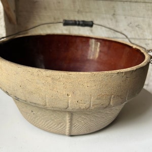 Puede incluir: Un tarro de barro vintage con un interior esmaltado marrón y un asa de alambre con un agarre de madera. El tarro tiene un exterior texturizado con un patrón decorativo.