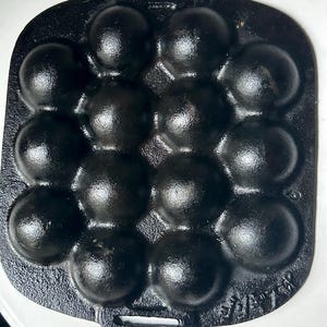 May include: A black cast iron pan with 14 spherical indentations for cooking. The pan has a rectangular shape with a handle on one side. The surface of the pan has a textured appearance, and the overall design is simple and functional.