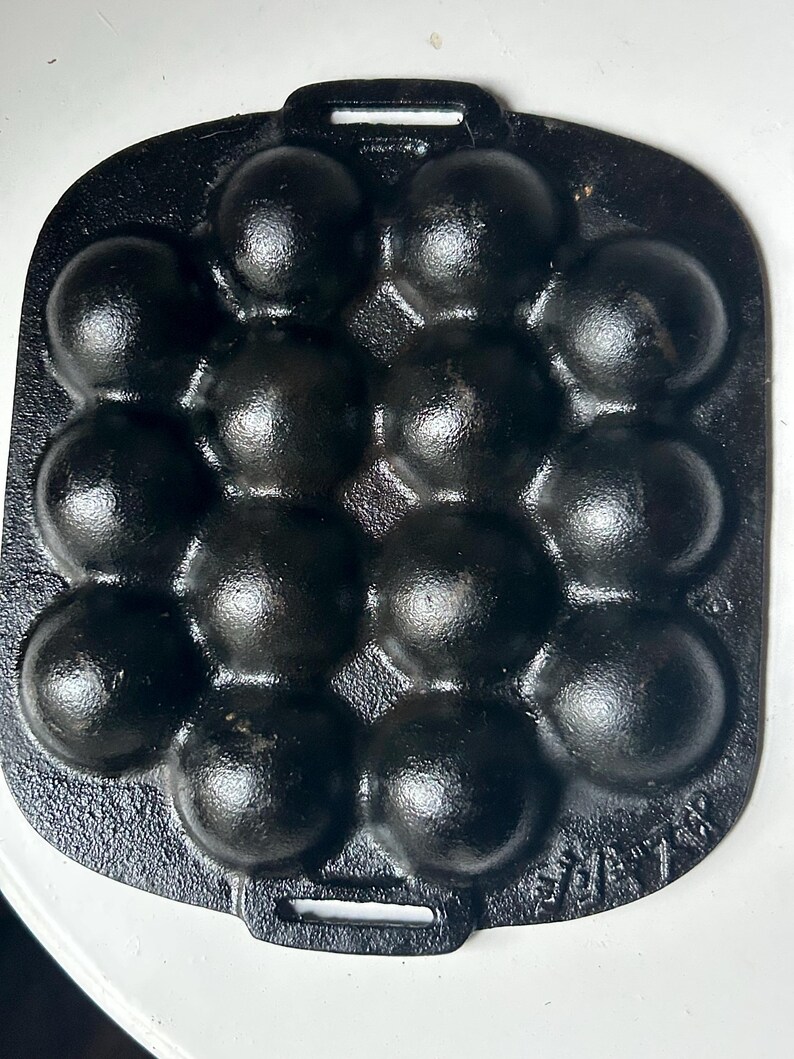 May include: A black cast iron pan with 14 spherical indentations for cooking. The pan has a rectangular shape with a handle on one side. The surface of the pan has a textured appearance, and the overall design is simple and functional.