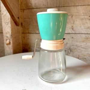 Op de afbeelding: Vintage handmolen voor de keuken met een heldere glazen pot, een turquoise bovenkant en een witte handgreep. De molen heeft een metalen slinger en een witte basis. De pot is ongeveer 15 cm hoog.