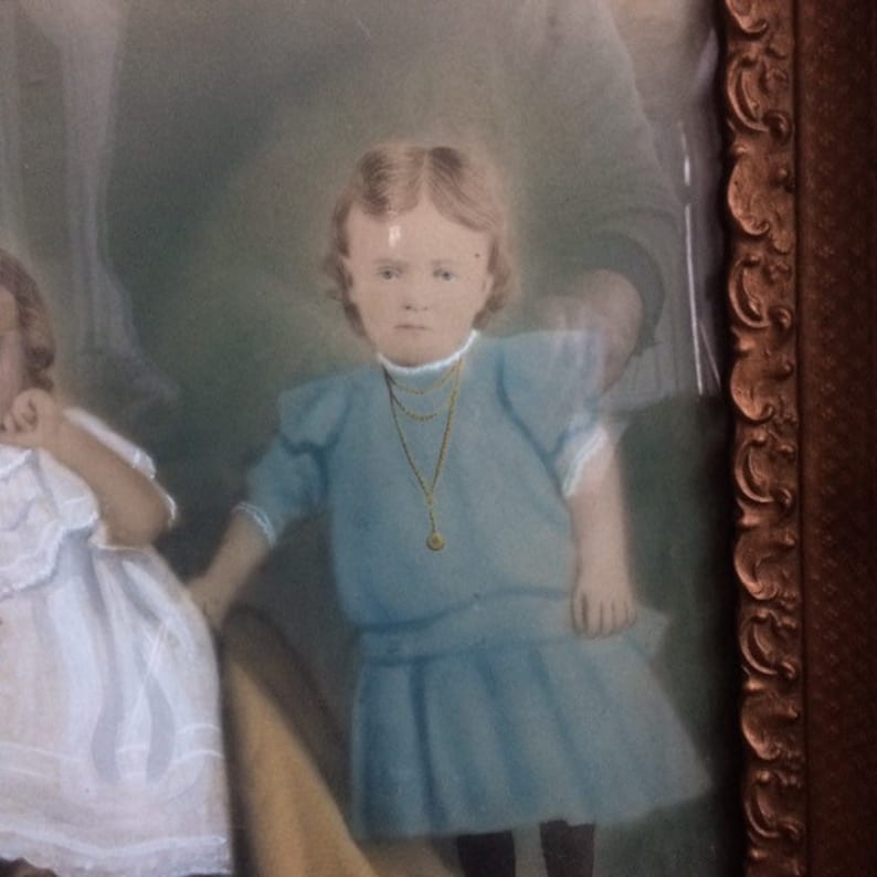 May include: A vintage photograph of two young girls in a frame. The girl in the foreground is wearing a blue dress with a gold necklace and a locket. The girl in the background is wearing a white dress.