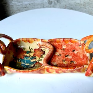 May include: A ceramic serving dish with two compartments and two loop handles. The dish is painted with a floral design in shades of red, orange, blue, and green. The handles are a bright orange color.
