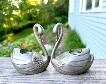 May include: A pair of silver-toned metal swan-shaped candle holders. Each swan features a curved neck, detailed wing feathers, and a circular opening on the back designed to hold a candle. The metal has a matte finish with visible texture.