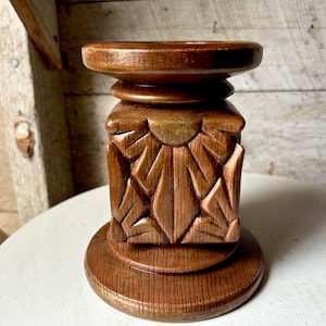 May include: A wooden candle holder with a carved design. The holder has a square base and a round top. The wood is a dark brown color.