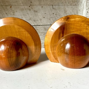 May include: Two wooden decorative objects. Each has a large, light-brown, semi-circular base with a smaller, darker-brown, spherical element in front. The wood has a glossy finish. The objects are displayed on a white surface.