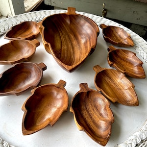 Könnte beinhalten: Ein Set aus sieben Holzschalen in Form von Blättern. Die Schalen sind aus dunkelbraunem Holz gefertigt und haben eine natürliche, rustikale Oberfläche. Die Schalen sind in einem Kreis auf einer weißen Oberfläche angeordnet.