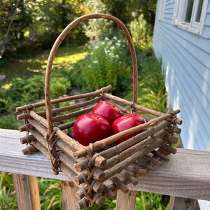 May include: A small, rustic basket made of woven twigs with a handle. The basket is filled with three red apples.