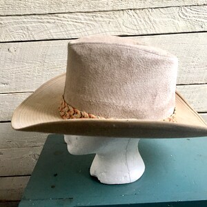 May include: A beige cowboy hat with a patterned band. The hat is on a mannequin head and is set against a white wood background.