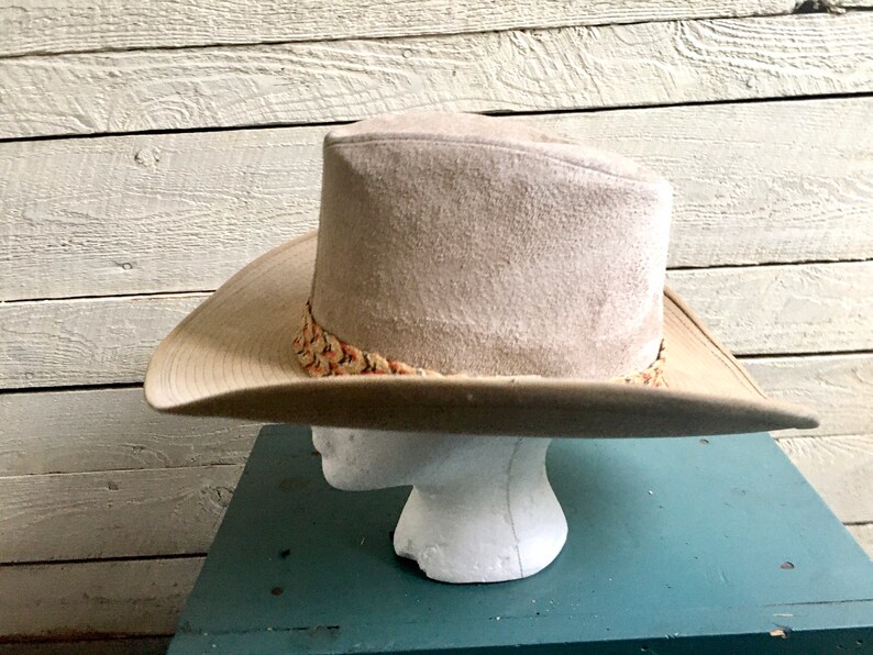 May include: A beige cowboy hat with a patterned band. The hat is on a mannequin head and is set against a white wood background.