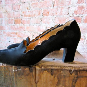 May include: Black suede heeled shoes with a scalloped edge and a low platform. The shoes are sitting on a wooden surface.