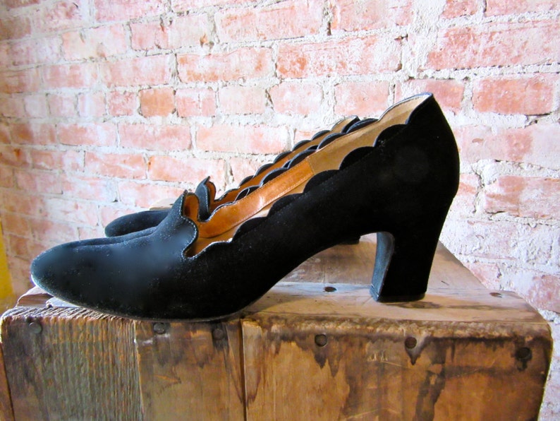 May include: Black suede heeled shoes with a scalloped edge and a low platform. The shoes are sitting on a wooden surface.
