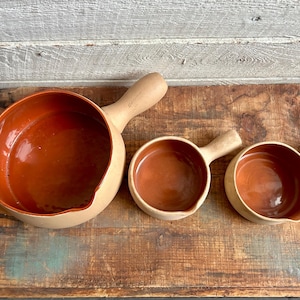 Könnte beinhalten: Set aus drei Keramik-Servierschalen mit Griffen. Die Schalen haben eine hellbeige Außenseite und eine warme braune Innenseite. Die Griffe sind aus hellem Holz gefertigt. Die Schalen sind auf einer rustikalen Holzoberfläche angeordnet.