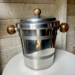 Può includere: Un secchiello per il ghiaccio in argento con un coperchio e manici in legno. Il secchiello ha una finitura lucida e lucidata ed è decorato con linee orizzontali.