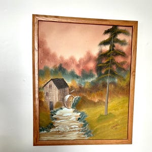 May include: A framed painting depicting a rustic watermill with a wooden water wheel, a stream, and a tall pine tree in the foreground. The background features a soft pink and green landscape with a hazy sky. The painting is signed "Chilens 95" in the bottom right corner.