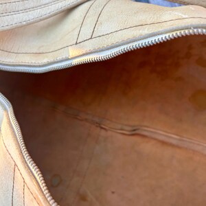 May include: A tan leather duffel bag with a silver zipper and a matching handle. The bag has visible stitching and a worn appearance, suggesting it is vintage or well-used. The interior of the bag is visible, showing a light brown color.