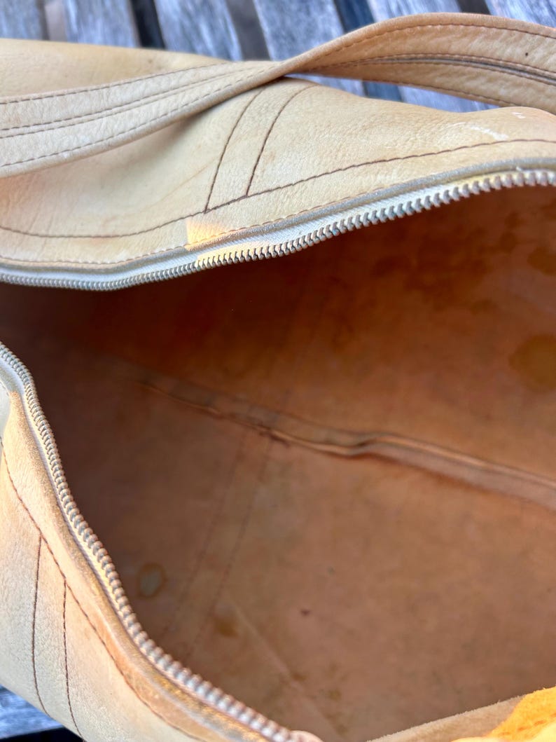 May include: A tan leather duffel bag with a silver zipper and a matching handle. The bag has visible stitching and a worn appearance, suggesting it is vintage or well-used. The interior of the bag is visible, showing a light brown color.