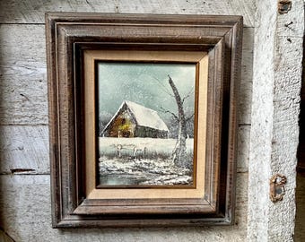 May include: A framed painting of a snowy winter scene. The artwork depicts a barn with a snow-covered roof, set against a backdrop of falling snow and bare trees. The frame is a dark, rustic brown.