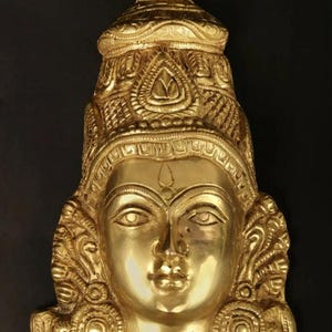 May include: A polished brass wall plaque featuring the sculpted face of a deity. The figure wears an ornate, tiered crown with intricate engravings and circular ear ornaments. The facial features are smooth with a central forehead mark.