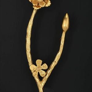 May include: A gold-toned brooch shaped like a Y-split branch featuring a textured, blooming flower at the top, a smaller five-petaled flower on the left stem, and a smooth, pointed bud on the right stem, all rendered in a metallic gold finish.