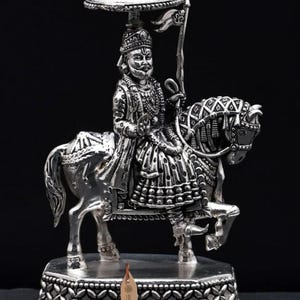 May include: A silver-toned metallic figurine depicting a royal figure riding a horse. The figure wears traditional attire, holds a flag, and is sheltered by an umbrella. The sculpture stands on an octagonal base with a decorative pattern and a small tag.