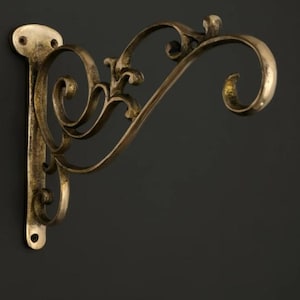 Puede incluir: Un soporte de pared de metal con acabado en latón oscuro, que presenta una placa de montaje vertical alargada con dos orificios para tornillos y un brazo curvo en forma de voluta que se extiende para colgar objetos con un gancho decorativo.