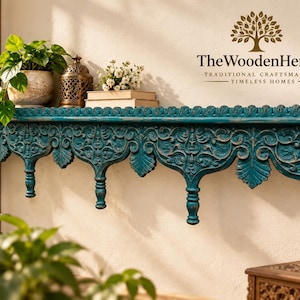 Puede incluir: Un estante de pared de madera color verde azulado con intrincados patrones florales tallados y remates colgantes. El estante está montado en una pared y muestra una planta en maceta, una linterna de metal y dos libros. El texto TheWoodenHeritage es visible.