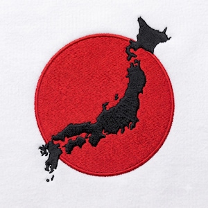 Puede incluir: Una superficie de tela blanca con un diseño bordado de un mapa negro sólido de Japón superpuesto sobre un gran parche circular rojo bordado. El bordado muestra un patrón de puntadas denso y texturizado en todo el círculo rojo y el mapa negro.