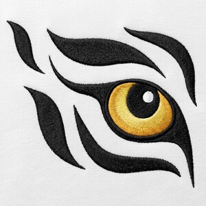 May include: An embroidered design on a white fabric surface featuring a stylized tiger eye. The eye has a bright yellow iris with a black pupil and a white highlight, surrounded by bold, sweeping black embroidered tiger stripe patterns.