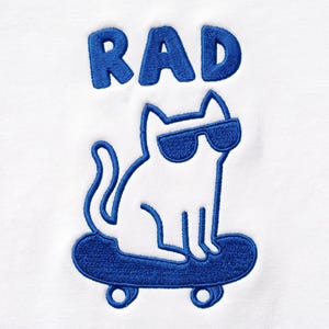 Può includere: Una superficie in tessuto bianco con un ricamo blu raffigurante un gatto con occhiali da sole su uno skateboard. Sopra il gatto, la parola RAD è ricamata in lettere maiuscole blu con un motivo a punto spesso e strutturato.