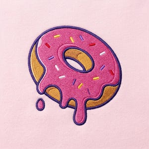 Puede incluir: Un diseño de rosquilla bordado sobre un fondo de tela rosa claro. La rosquilla presenta una base de color marrón dorado con glaseado rosa brillante que gotea por los lados, acentuado por pequeñas chispas multicolores en amarillo, blanco, azul y rojo.