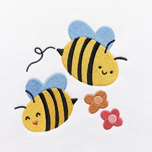 May include: An embroidery design on white fabric featuring two yellow and black striped bees with light blue wings. One bee has a curly flight path line, and both have simple smiling faces. Two small flowers, one orange and one pink, are below.