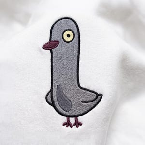 Puede incluir: Una prenda de tela blanca con una paloma gris bordada con un contorno oscuro. La paloma tiene un ojo amarillo pálido con pupila negra, un pico color granate oscuro y patas a juego. El ave está representada en un estilo sencillo y gráfico.